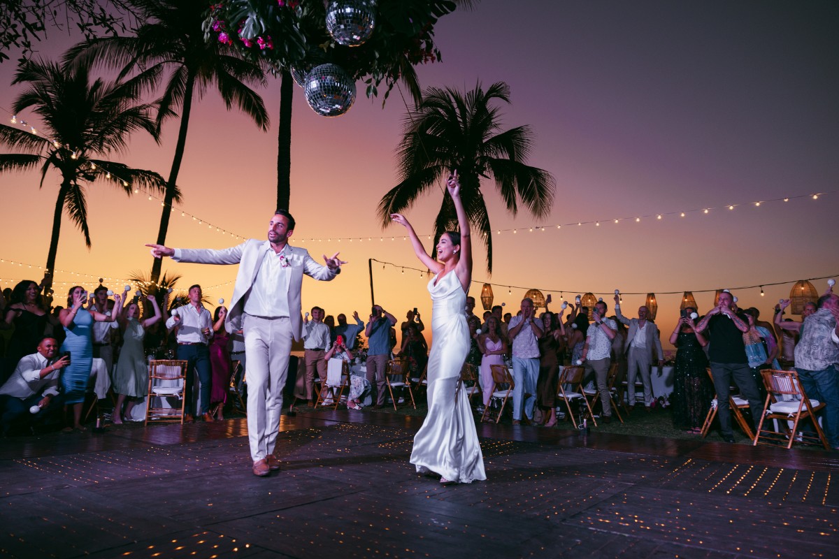 A group of people celebrates, including a bride in a white dress and a groom in a tan suit, posing together with arms raised at a festive event.