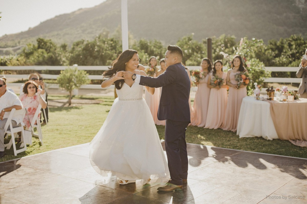 A bride and groom dance outdoors at a wedding reception with bridesmaids and guests watching, set against a backdrop of greenery and mountains.