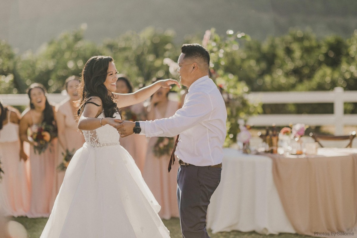 A bride and groom dance outdoors in front of a table with decorations. Bridesmaids in matching dresses watch in the background.