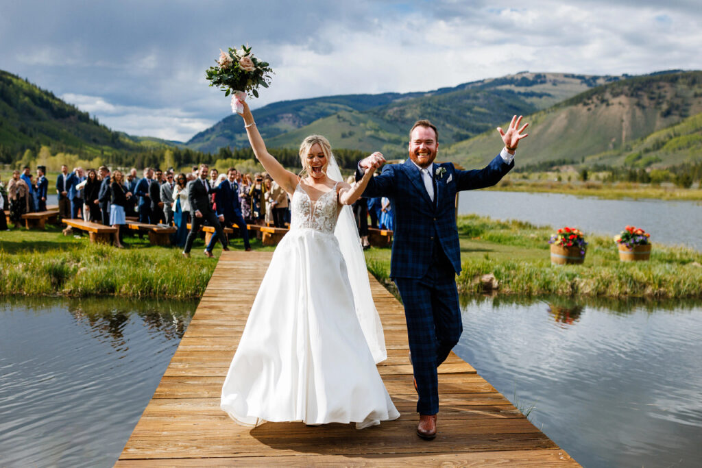 A bride and groom walk hand in hand down a wooden walkway over water, with a cheering crowd and mountainous landscape in the background.