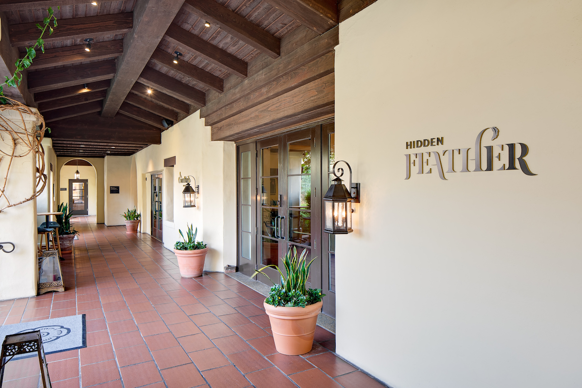 A covered outdoor walkway with terracotta tiles, two potted plants, and a door. A sign on the wall reads "Hidden Feather.