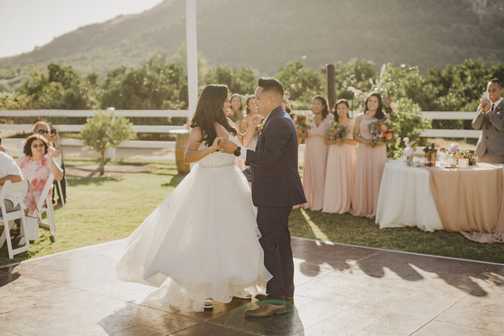 A bride and groom do their First Dance outdoors at their wedding reception, with bridesmaids in pink dresses watching in the background.