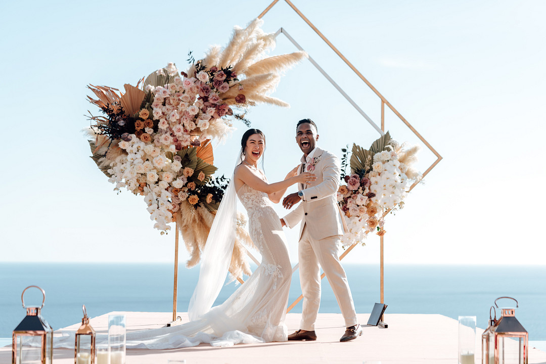 A couple stands at an altar adorned with flowers, exchanging vows under a canopy. Guests are seated on both sides, facing the ocean under a clear sky.