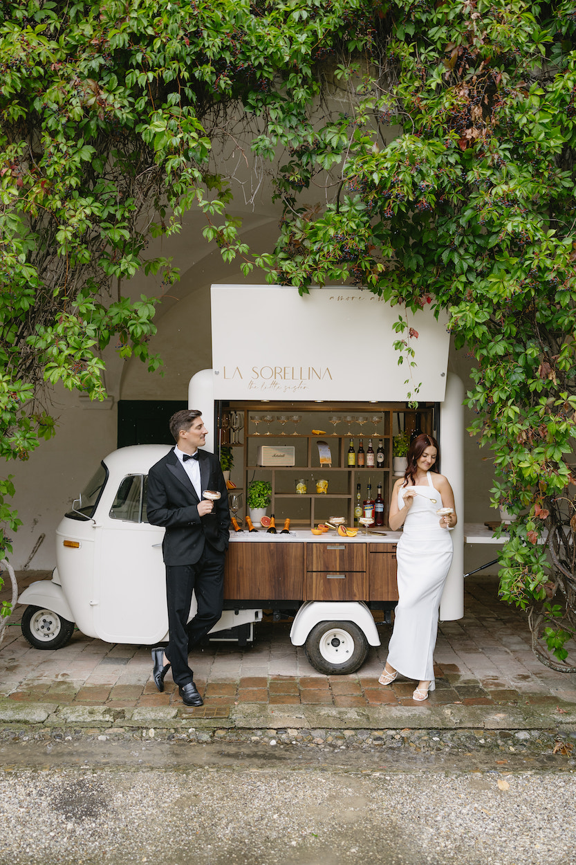 A man and woman in formal attire stand by a vintage mobile bar, adorned with greenery.