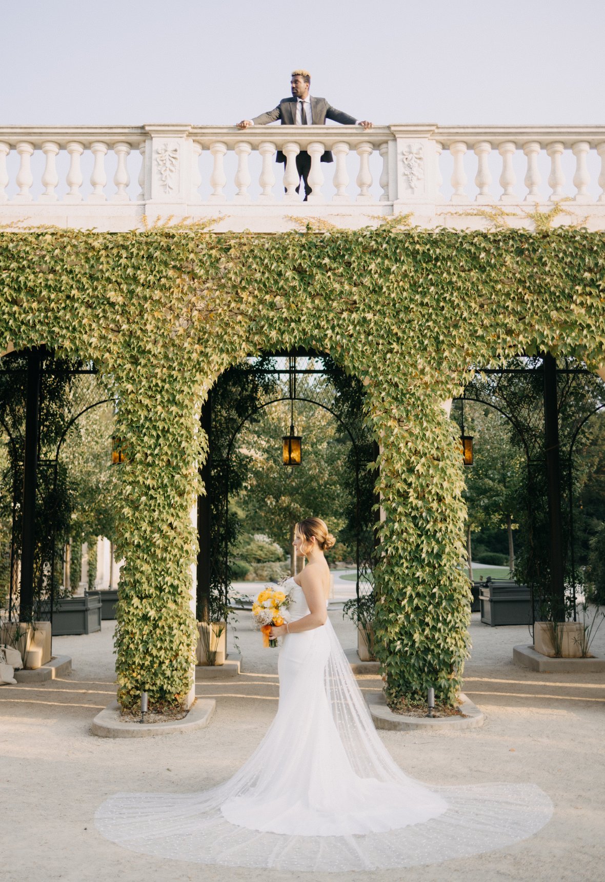 A bride in a white dress stands under a vine-covered arch, holding a bouquet. A person in a suit looks down from a balcony above.