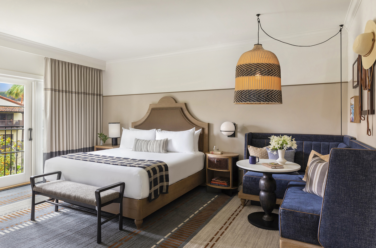 A cozy hotel room with a queen bed, a bench, a round table with blue seating, a large woven light fixture, and wall decor, including hats. A window with curtains is on the left.