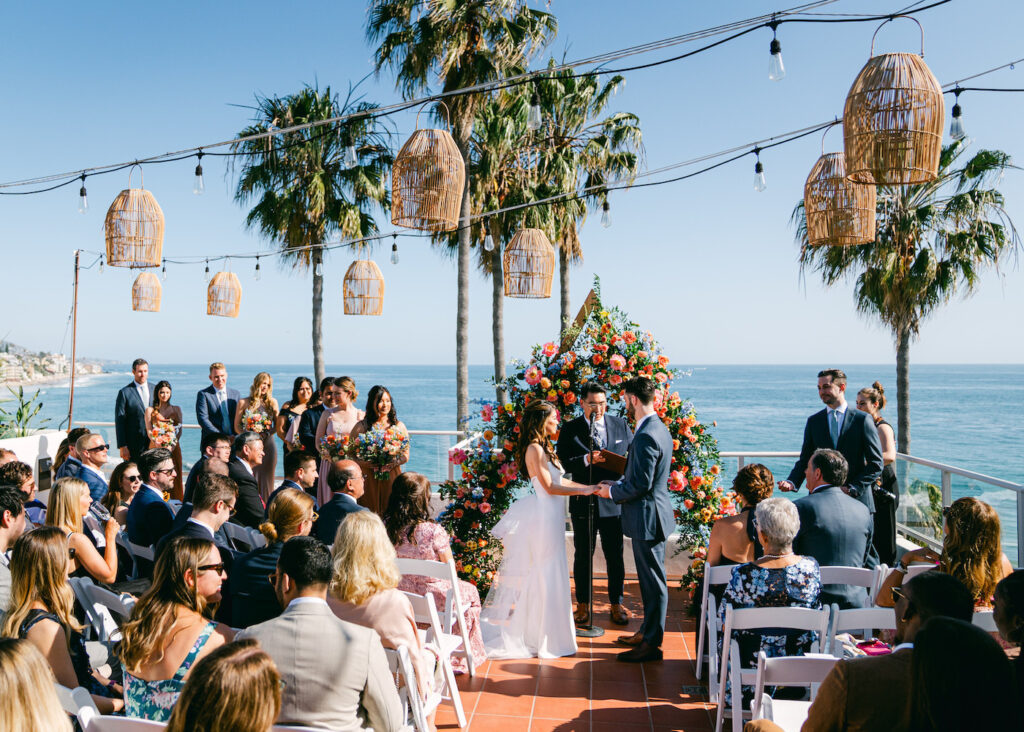 Outdoor wedding ceremony by the ocean, featuring a couple exchanging vows under a floral arch, surrounded by guests, palm trees, and decorative lights.