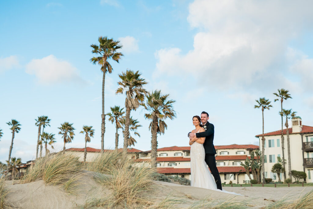 A couple stands on a sandy beach with palm trees and buildings in the background under a partly cloudy sky.