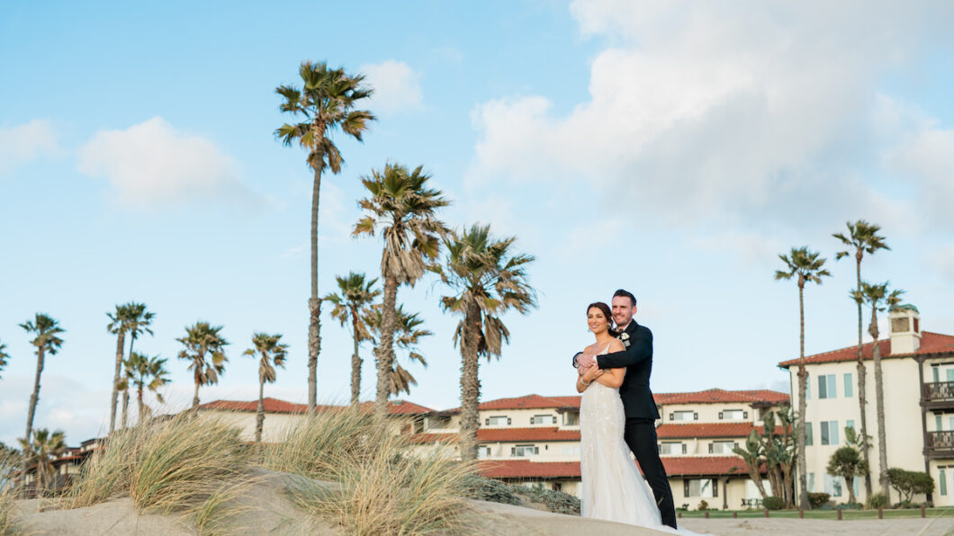 A couple stands on a sandy beach with palm trees and buildings in the background under a partly cloudy sky.