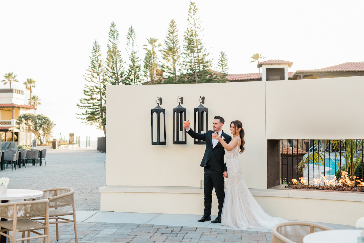 A couple in formal attire toasts outdoors near a wall with large lanterns. Trees and a building are in the background.