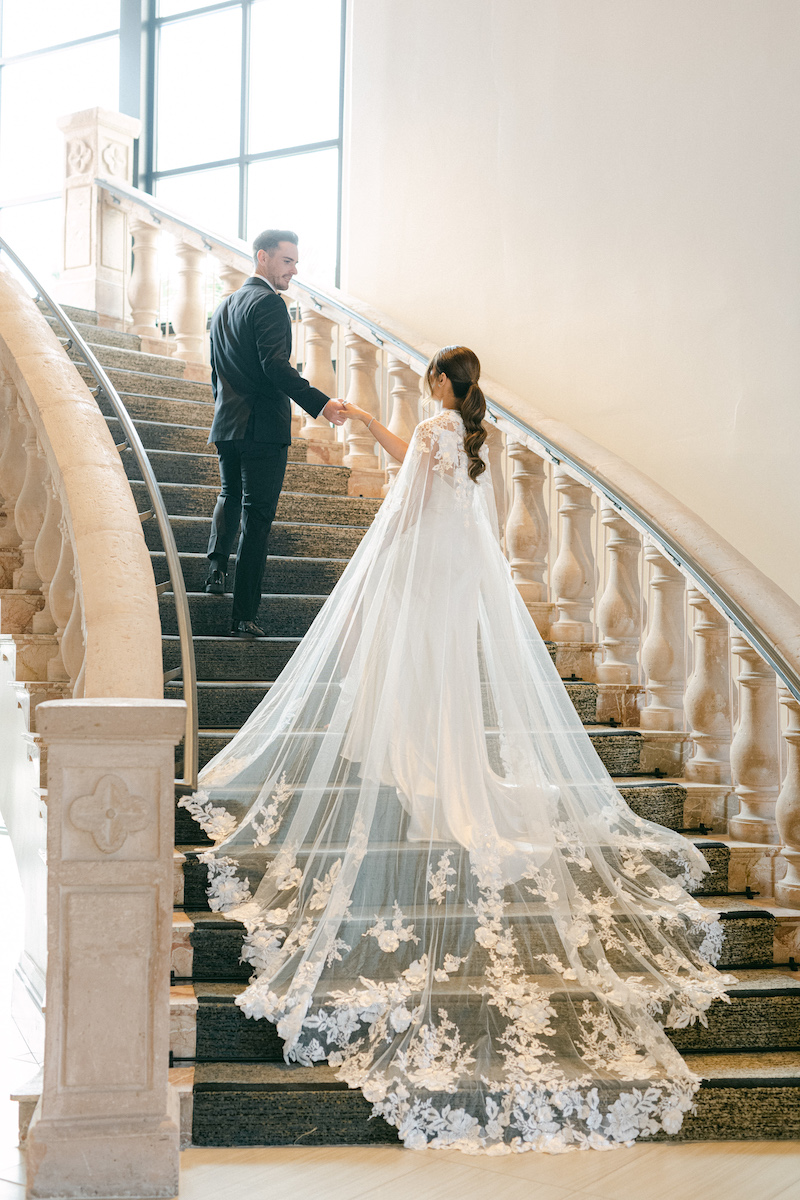 A bride and groom ascend a grand staircase. The bride wears a flowing wedding dress with an ornate train. The groom is in a black suit.