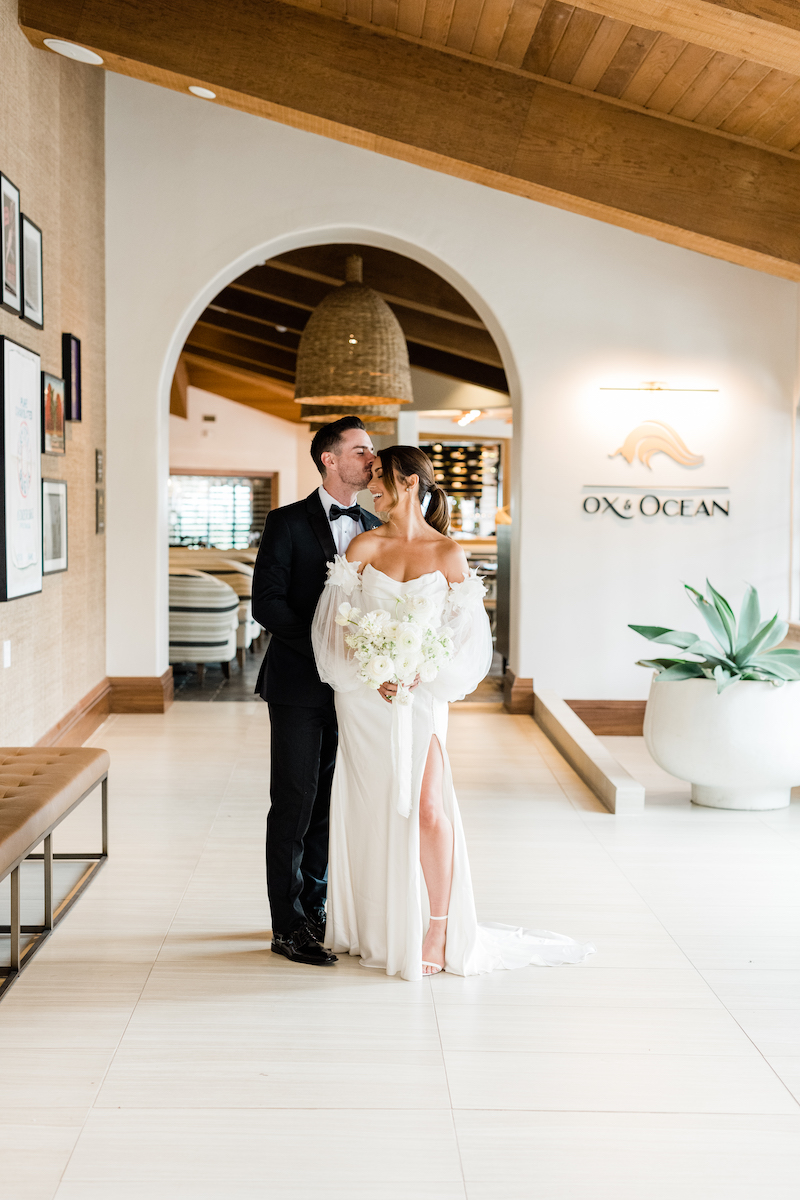 A bride and groom stand together in a bright, elegantly decorated hallway, with a sign reading "OX &amp; OCEAN" in the background. The groom is in a tuxedo; the bride holds a bouquet. 