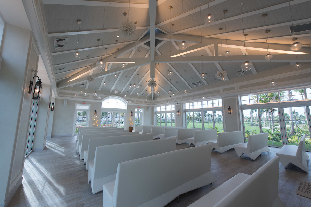 A modern room with a vaulted ceiling, white benches, and contemporary light fixtures. Large windows offer views of greenery outside.