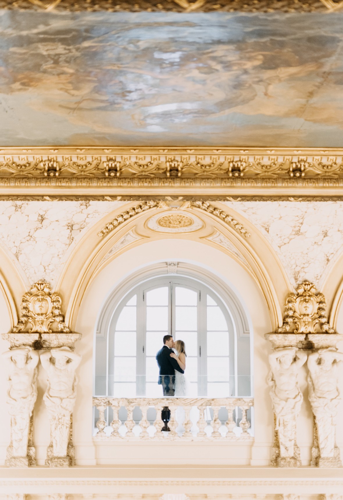 A couple embraces in a light-filled arched balcony, surrounded by ornate architecture and a decorative ceiling.