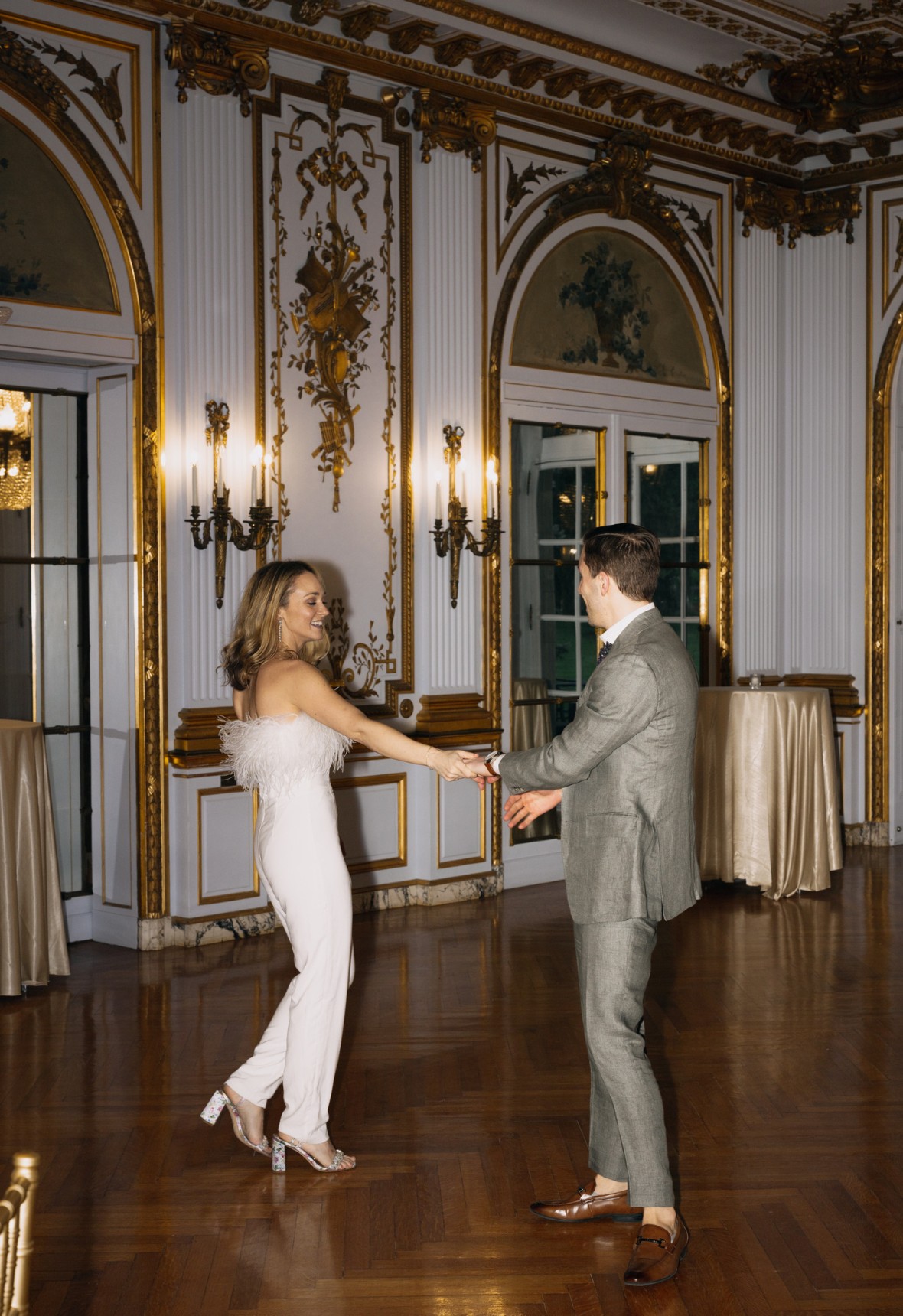 A couple dances in an elegant room with ornate gold details, wood flooring, and large arched windows. The woman wears a white outfit, and the man wears a gray suit.