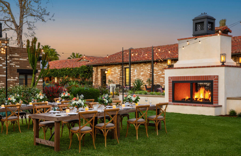 Outdoor dining setup with wooden tables and chairs on a lawn, a lit fireplace nearby, and a brick building in the background at sunset.