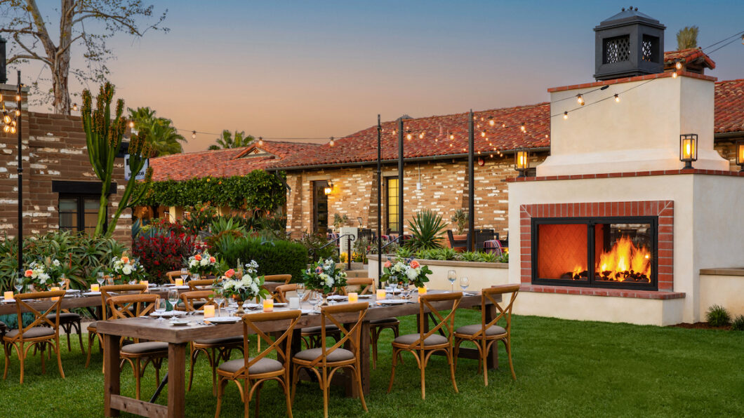 Outdoor dining setup with wooden tables and chairs on a lawn, a lit fireplace nearby, and a brick building in the background at sunset.