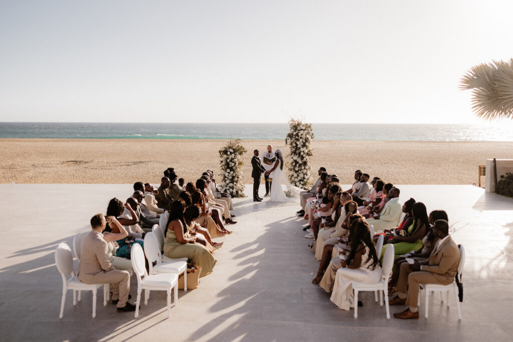 A beach wedding ceremony with a couple standing under floral arches. Guests are seated on either side of the aisle, facing the ocean.