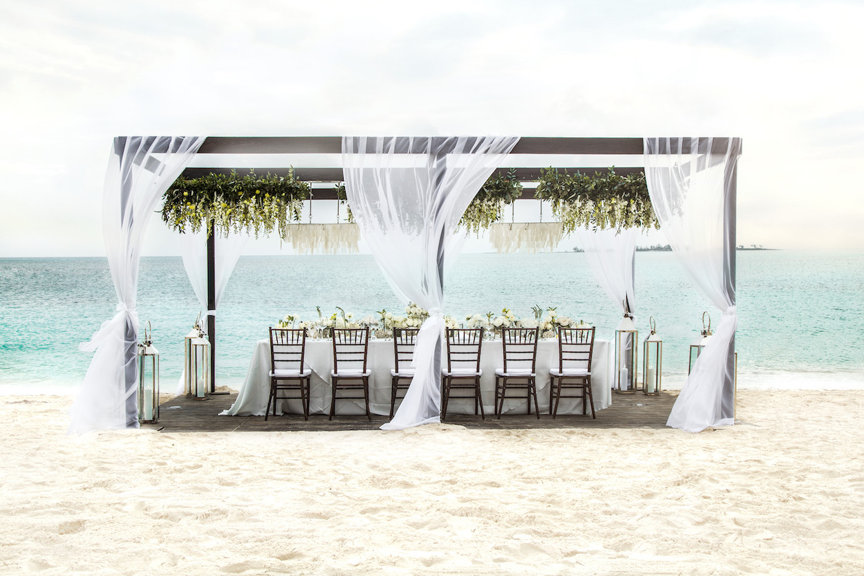 Outdoor beach wedding setup with a long table under a canopy draped with white curtains and greenery.