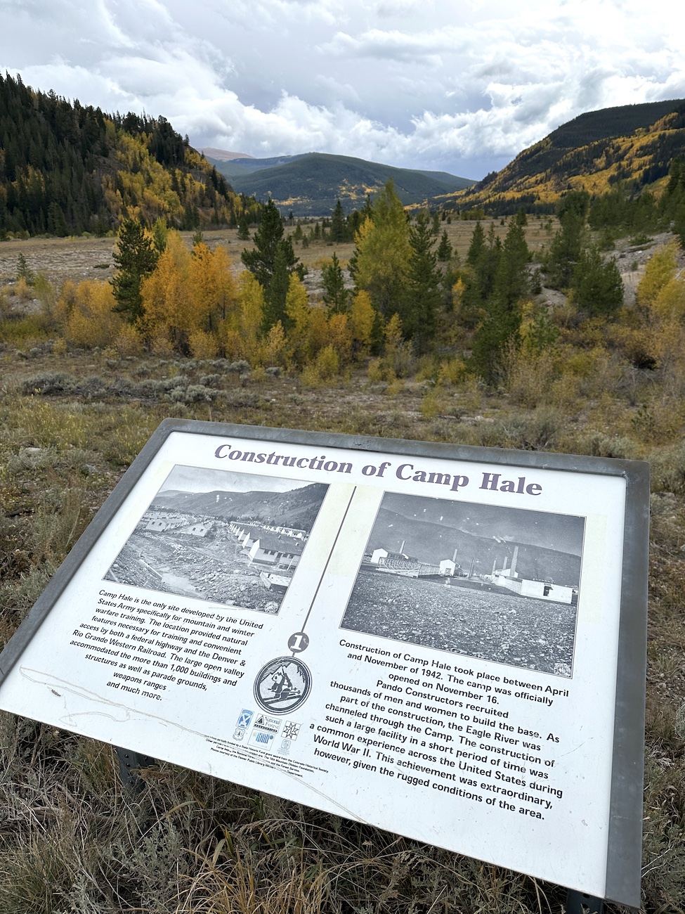 Informational sign about Camp Hale in a mountainous landscape with cloudy sky, surrounded by trees with yellow and green foliage.