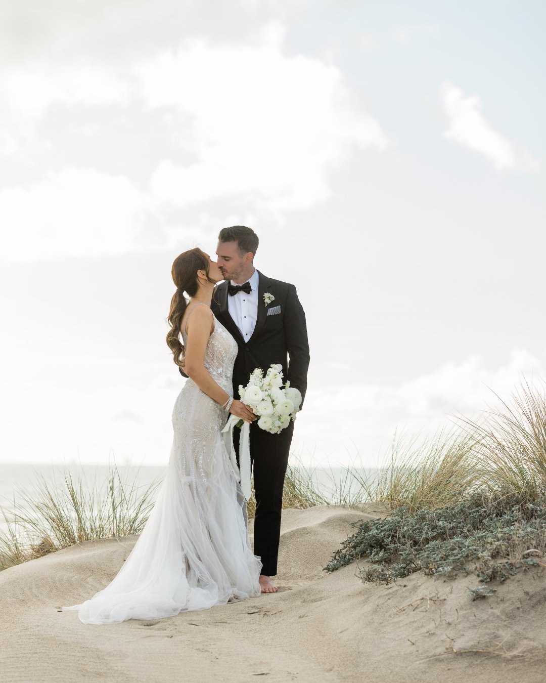 Bride and groom stand on sand dunes, embracing and holding a bouquet, with ocean and sky in the background.