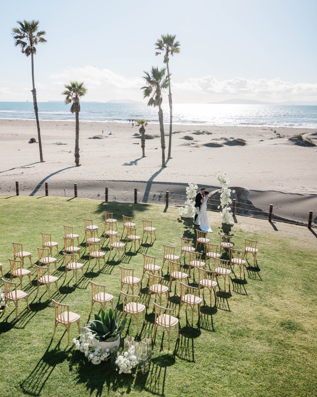 Outdoor beach wedding setup with rows of chairs on grass facing an ocean view. Two people standing near a floral arch under palm trees.