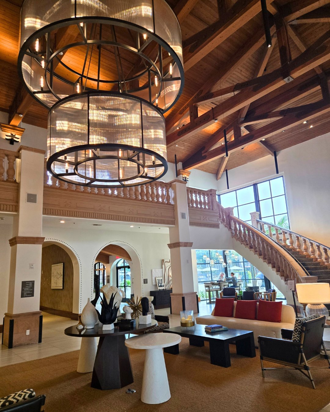 Spacious lobby with high ceilings, large circular chandeliers, wooden beams, a grand staircase, and modern furniture. Natural light streams through large windows.