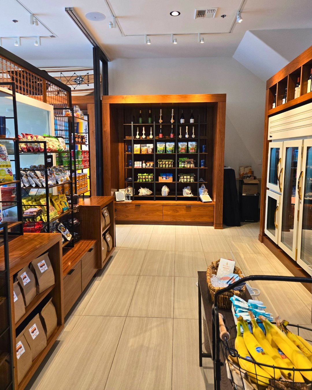 A small market displays shelves with snacks, beverages, and condiments. A basket of bananas sits near refrigerated sections. Warm lighting enhances the inviting atmosphere.