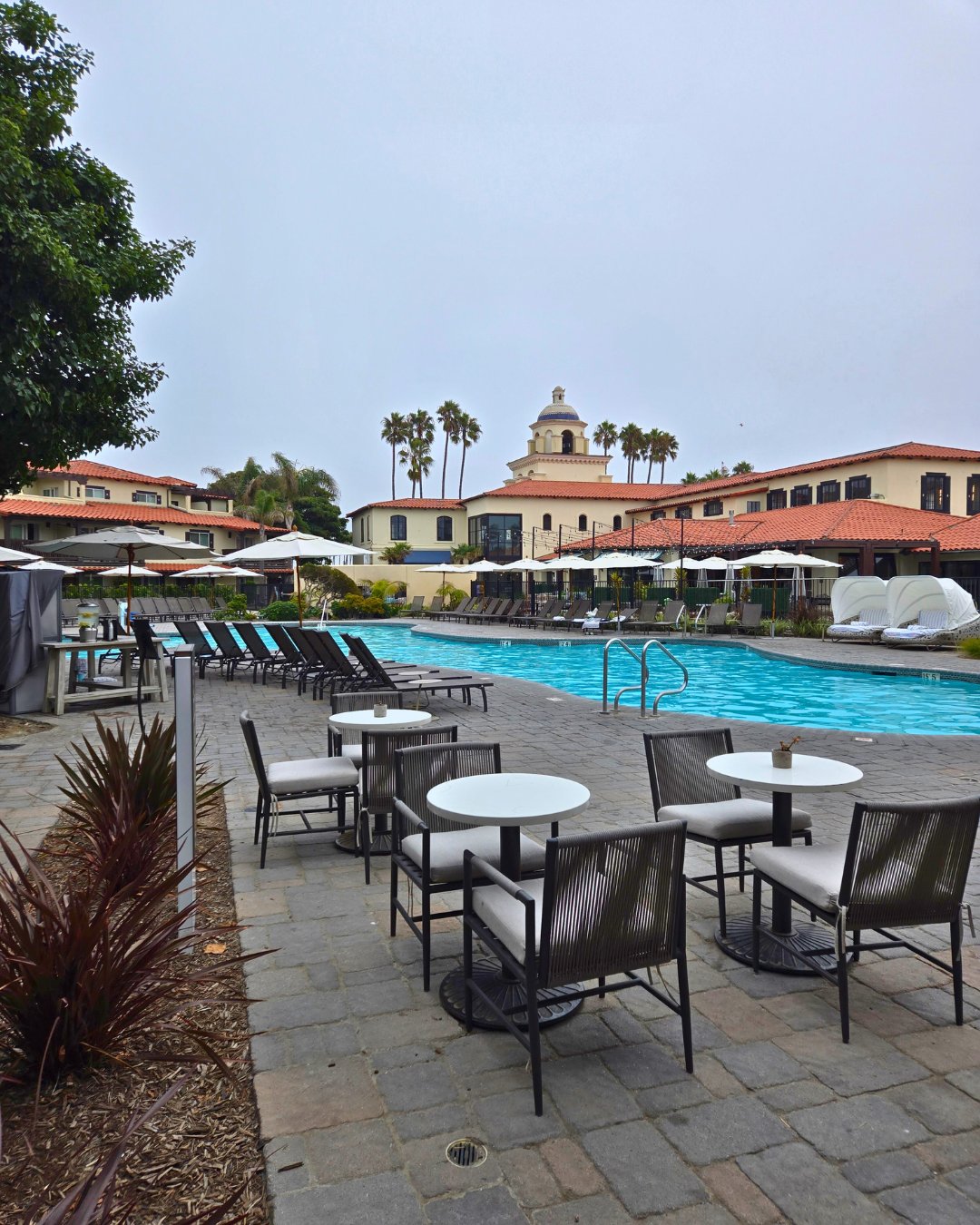 Outdoor pool area at a resort with lounge chairs and tables. The building in the background has a dome and palm trees surround the scene.