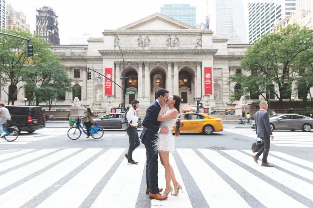 A couple kisses on a crosswalk in front of an ornate building, with pedestrians, cyclists, and a yellow taxi nearby.