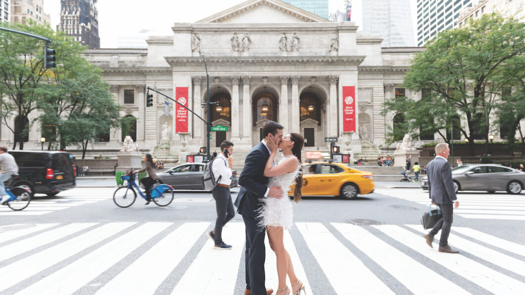 A couple kisses on a crosswalk in front of an ornate building, with pedestrians, cyclists, and a yellow taxi nearby.