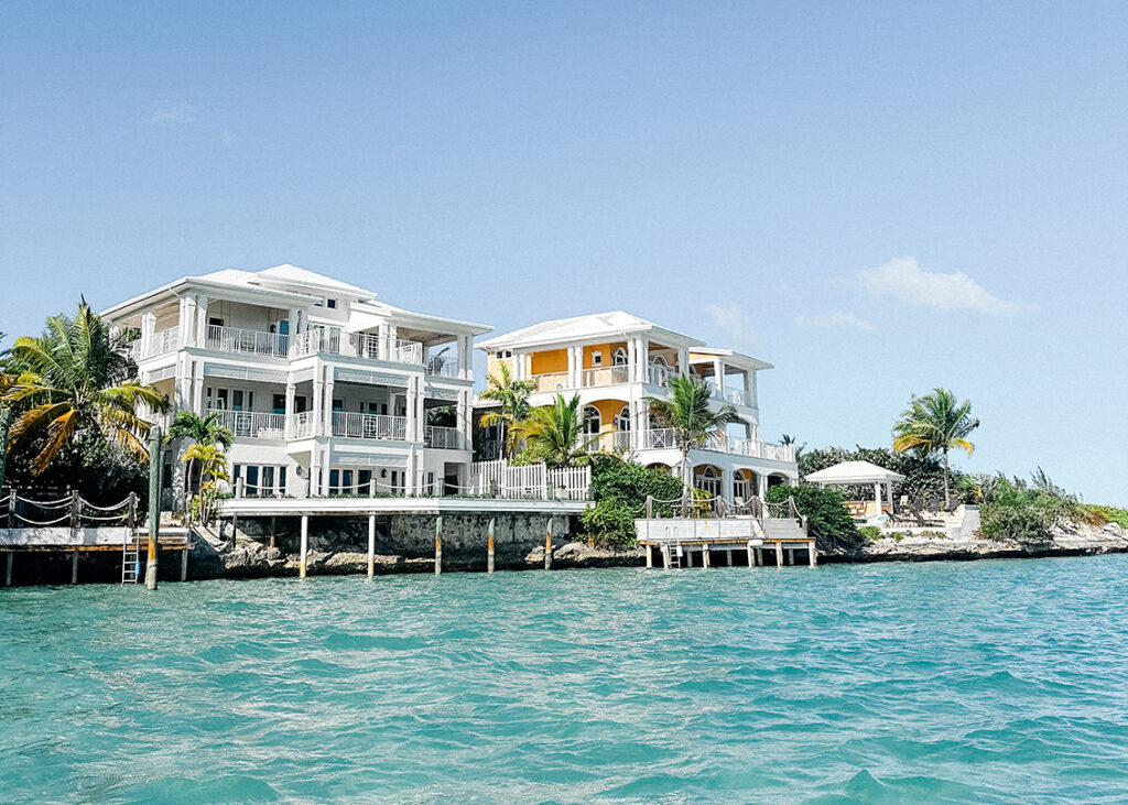 Oceanfront houses with white and yellow facades, surrounded by palm trees, along a turquoise shoreline under a clear blue sky.