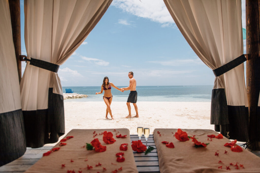 A couple holds hands on a sandy beach, viewed from a cabana with curtains. Red flowers and towels are on a table in the foreground.