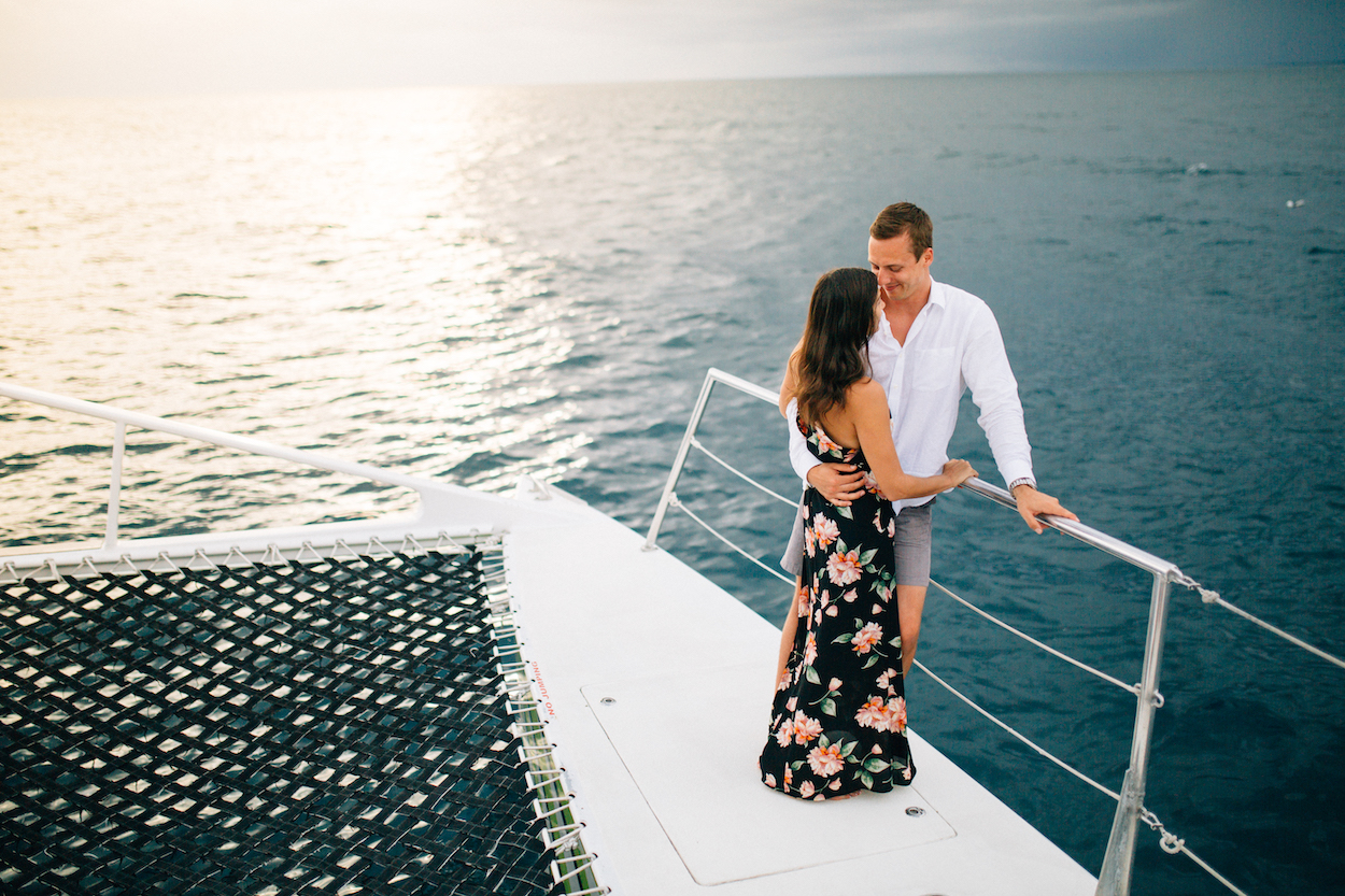 A couple stands on a boat at sunset, embracing near the railing and looking at each other.