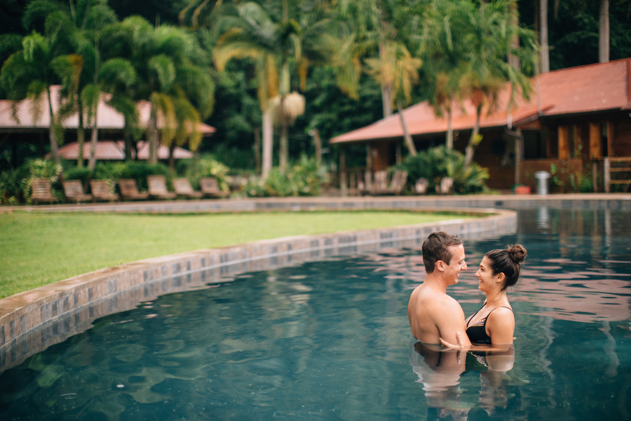 A couple stands in a pool, facing each other and smiling. In the background are palm trees and wooden buildings.