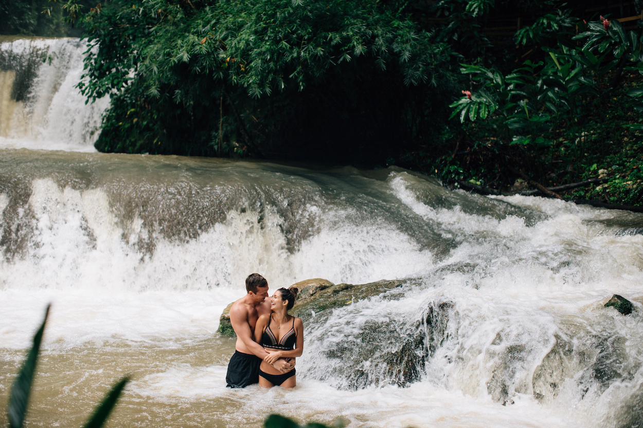 A couple stands in a river, in front of cascading waterfalls, surrounded by lush green foliage.