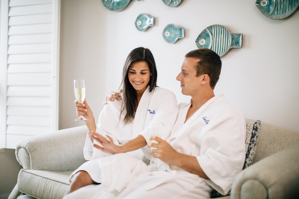 A couple in white robes sit on a couch, holding glasses of champagne and smiling. Wall has decorative fish art.