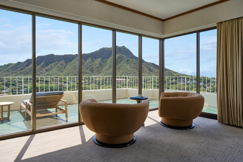 Hotel room with two curved chairs facing large windows, offering a view of a mountain landscape and a balcony with a lounge chair and small table.