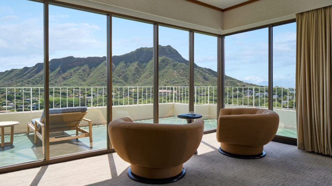 Hotel room with two curved chairs facing large windows, offering a view of a mountain landscape and a balcony with a lounge chair and small table.