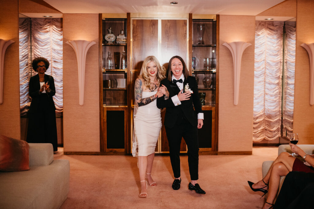 A couple in wedding attire enters a room, smiling. The person on the left wears a dress; the one on the right wears a tuxedo. An attendee applauds in the background.