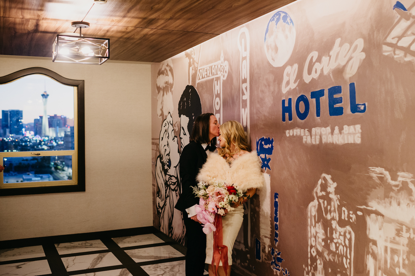 A couple in formal attire posing in a room with vintage-style Las Vegas artwork. One person is holding a bouquet, and a city view is visible through the window.