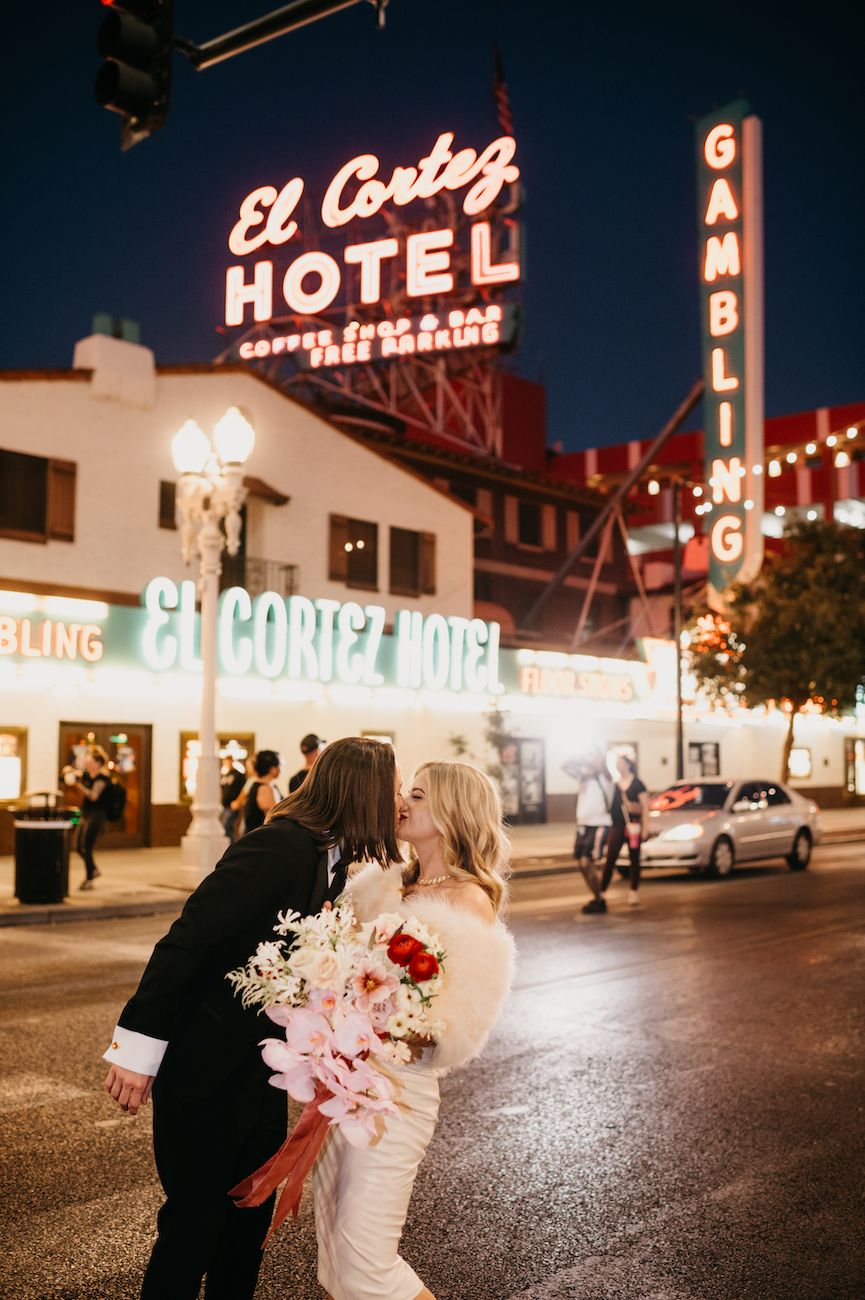 A couple in formal attire kisses on a street at night, under the illuminated sign of El Cortez Hotel. The woman holds a bouquet of flowers.