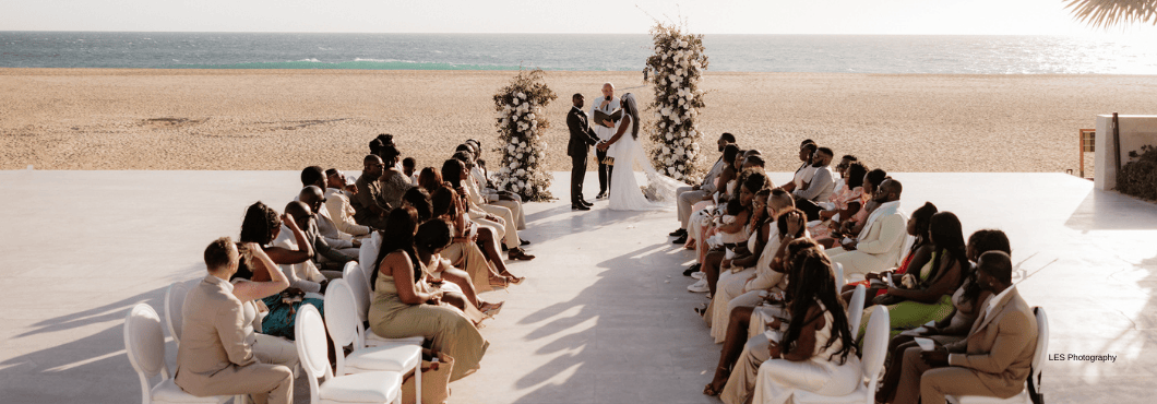 A beachside wedding ceremony with a couple under a floral arch, surrounded by seated guests.