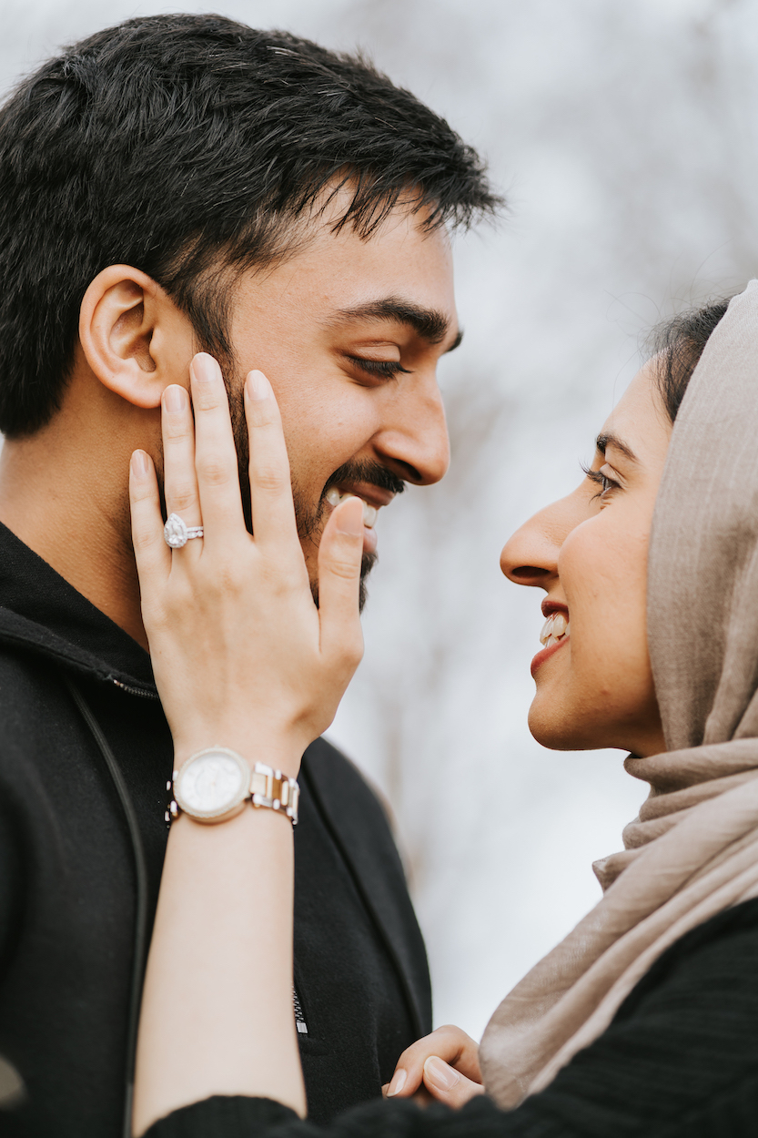 A couple smiling at each other; the woman touches the man's face, showcasing an engagement ring.