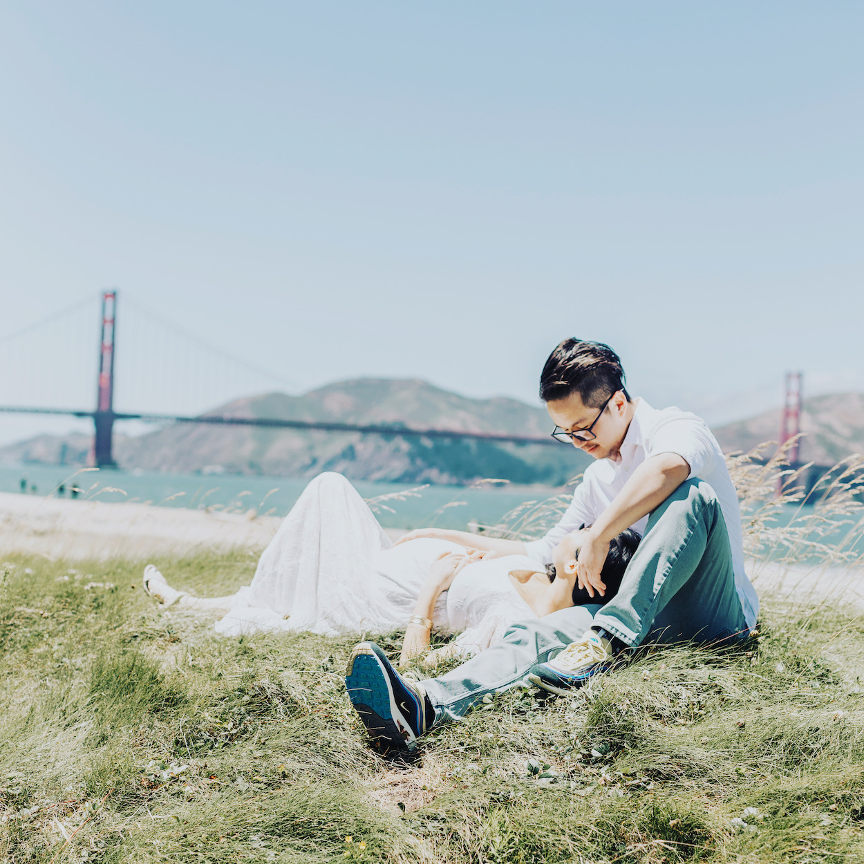 A couple relaxes on grassy ground near a beach with the Golden Gate Bridge in the background on a sunny day.