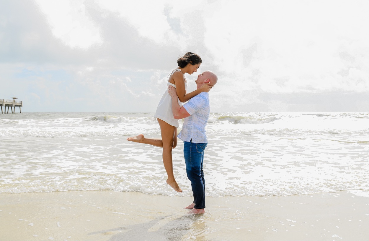 A man lifts a woman on a sunny beach with gentle waves in the background.