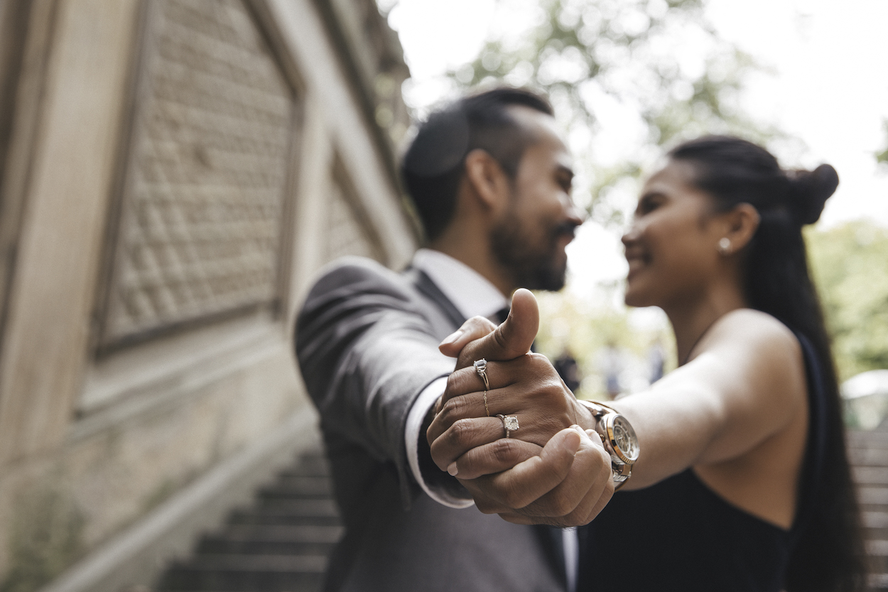 A couple holding hands in focus, wearing formal attire with visible rings, standing on outdoor steps. Trees and a stone wall are in the background.