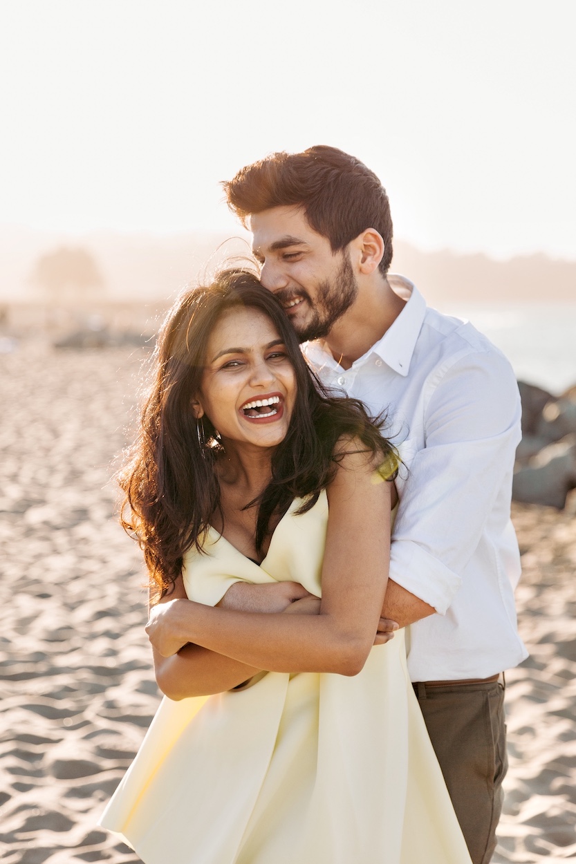 A couple laughing and embracing on a sunny beach with sand and blurred background.