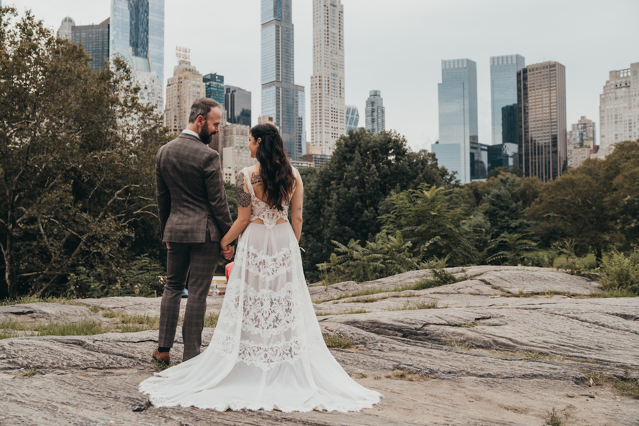 A couple in wedding attire stands on a rocky path, holding hands, with a city skyline and trees in the background.