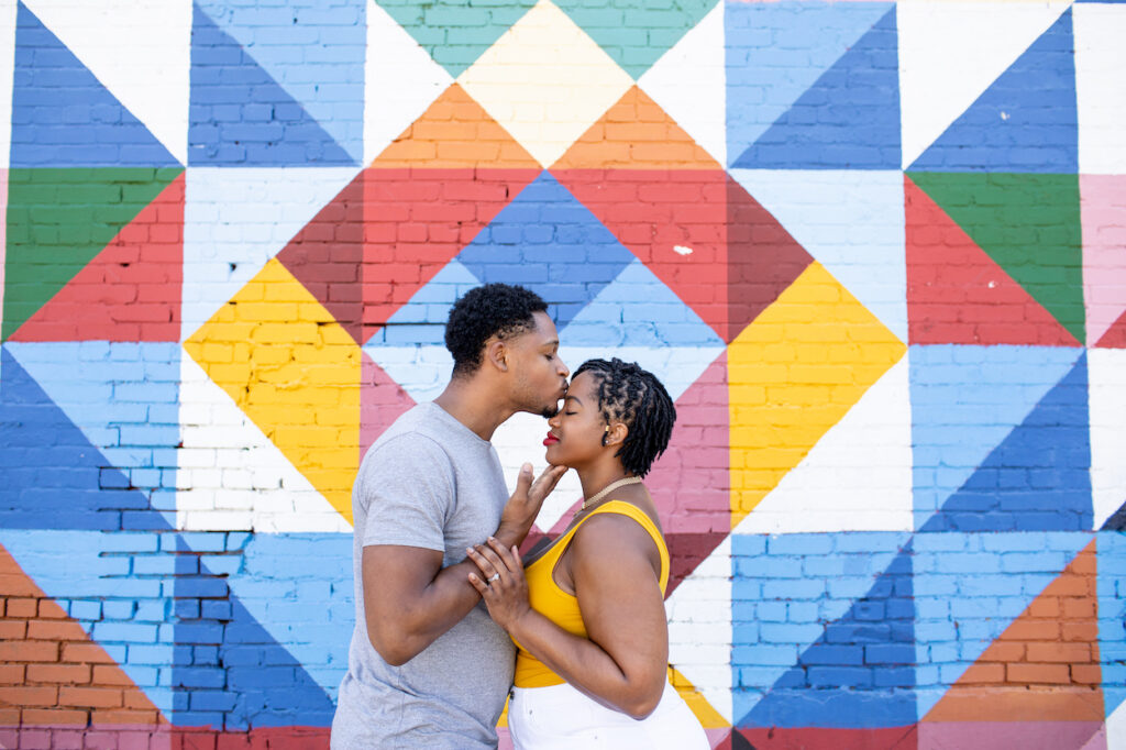 A couple stands in front of a colorful geometric mural. The man kisses the woman's forehead as they hold hands.